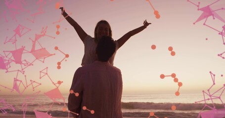Man lifting woman in casual beachwear on sandy shore during sunset, with geometric node overlay