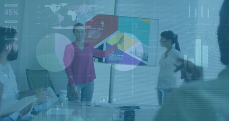 Presenting woman wearing purple blouse pointing toward data screen in boardroom, with tablet laptop
