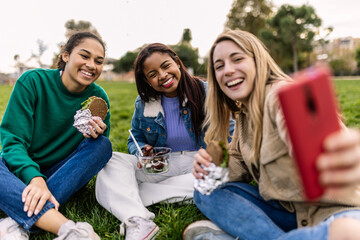 Diverse young women friends having social picnic in park, laughing and taking selfie with smartphone, enjoying outdoors lunch