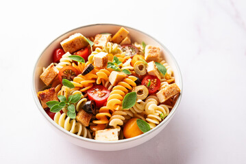 Warm salad with colorful full grain fusilli pasta, olives, fried tofu, cherry tomatoes and aromatic oregano leaves in ceramic bowl on white marble table top view. Healthy vegan food concept.