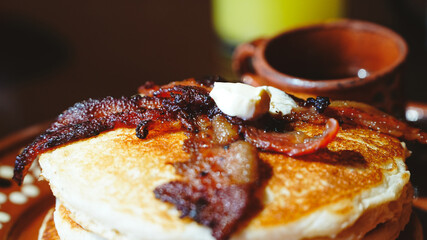 Plato de barro con una torre de hotcakes decorados con tocino y mantequilla, acompañado de una pequeña taza artesanal. La luz cálida resalta la textura esponjosa y el tono dorado del desayuno.