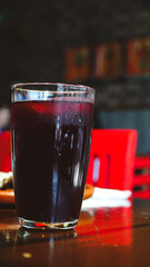 Vaso alto con bebida de color oscuro sobre una mesa de madera, con fondo difuminado y sillas rojas que aportan contraste.