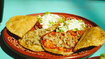 Platillo mexicano con antojitos variados —sopes, tacos dorados y gorditas— servidos en plato de barro sobre una mesa turquesa con luz natural.