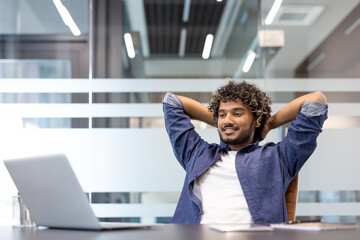 Happy young Indian man is reading in the office at a table in front of a laptop, hands behind his...