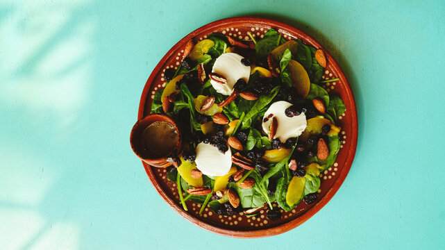Ensalada fresca servida en un plato de barro, con mezcla de espinacas, aguacate, almendras, pasas y aderezo en una peque&ntilde;a cazuelita. Los tonos verdes y dorados contrastan con la mesa turquesa
