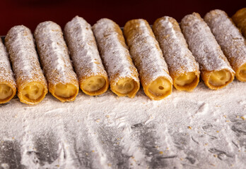 Row of cream-filled pastry rolls covered with powdered sugar on metal tray. Sweet dessert close-up.