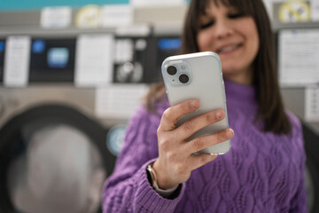 Young woman using smartphone in laundromat while waiting for laundry