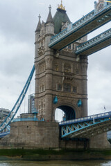 Naklejka premium Tower Bridge's ornate Gothic tower and blue supports, rising from the Thames under a cloudy sky.