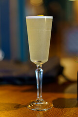 Pale yellow cocktail in a tall stemmed glass with a citrus garnish, sitting on a wooden table against a soft, dark, blurred background.