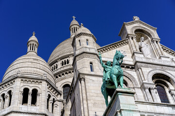 Upward view of Sacré-Cœur Basilica's white domes, intricate architecture, and green equestrian statue against a clear blue sky.