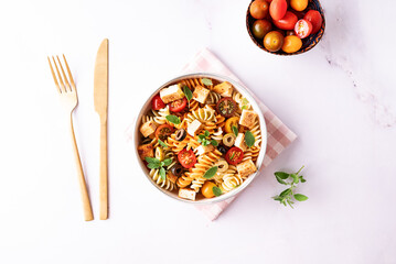 Warm salad with colorful full grain fusilli pasta, olives, fried tofu, cherry tomatoes and aromatic oregano leaves in ceramic bowl on white marble table top view. Healthy vegan food concept.