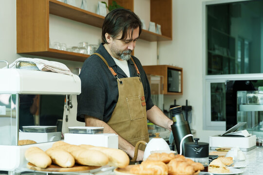 Senior Caucasian male barista wearing brown apron attentively using point-of-sale machine inside cozy small cafe handling customer transactions professionally., Concept small business
