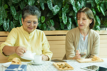 Asian senior woman pours tea while Caucasian woman smiles at a cafe table, enjoying a joyful and...