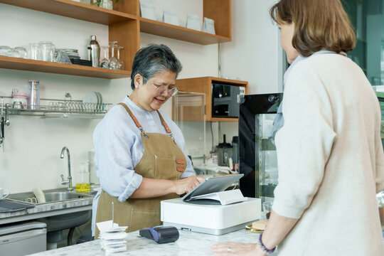 Asian senior female barista adult interacting with customer at cafe counter using modern point-of-sale system reflecting friendly atmosphere in small family coffee shop business - Powered by Adobe
