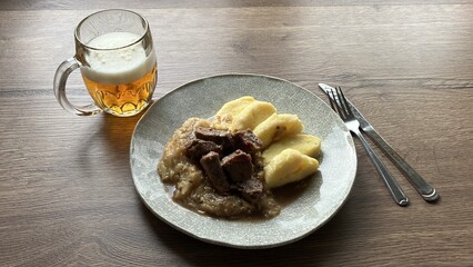 Traditional Czech dish of roasted wild boar with sauerkraut and potato dumplings, served on a wooden table with a glass of beer in the background. A comforting autumn meal representing rustic