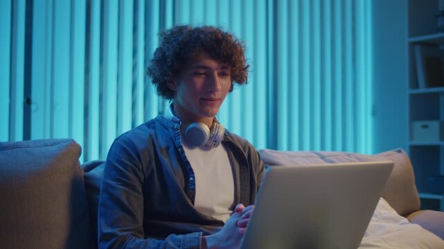 Young Man Sits on Sofa at Night with Laptop on Lap, Attending an Online Meeting. Casual Yet Productive Evening in a Hybrid Lifestyle. Remote Work, Digital Communication, Focus Mode.