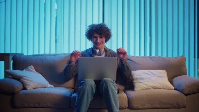 Young Man Sits on Sofa at Night with Laptop on Lap, Attending an Online Meeting. Casual Yet Productive Evening in a Hybrid Lifestyle. Remote Work, Digital Communication, Focus Mode.