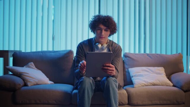Young Man Sits on Sofa at Night with Laptop on Lap, Attending an Online Meeting. Casual Yet Productive Evening in a Hybrid Lifestyle. Remote Work, Digital Communication, Focus Mode.