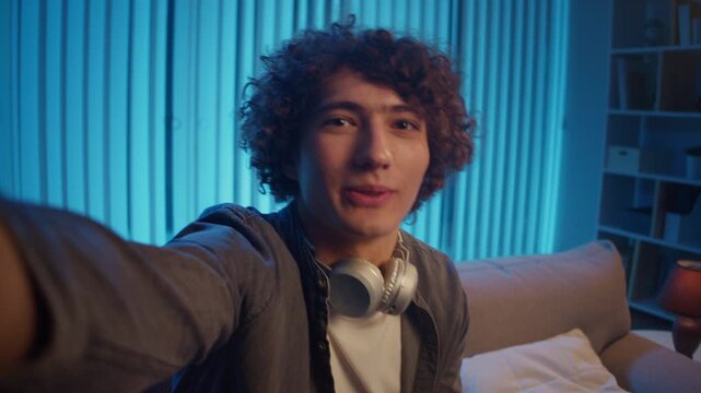 Young Man on Sofa Holds Phone at Night, Looking Into Camera During Video Call. Intimate Tech-Driven Moment in Remote Conversation. Virtual Presence, Digital Communication, Emotional Connection.