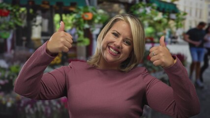 Young blonde woman wearing a pink sweater showing both thumbs up in front of a vibrant flower shop on a bustling street; approval.
