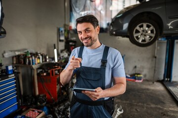 Confident mechanic pointing at camera and holding digital tablet in auto repair shop