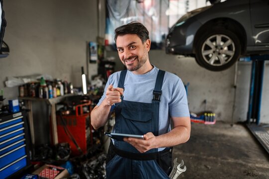 Confident mechanic pointing at camera and holding digital tablet in auto repair shop