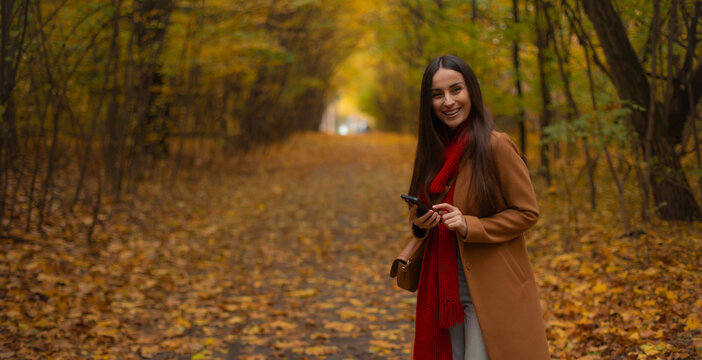 Smiling young woman with long hair using smartphone in autumn forest, wearing brown coat and red scarf, standing on a path covered with yellow leaves, enjoying fall day outdoors. - Powered by Adobe