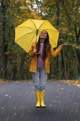 Obraz premium Happy young woman in yellow raincoat and rubber boots standing on rainy road with yellow umbrella, smiling and enjoying autumn rain in forest park.
