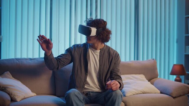 Young Man Sits on Sofa at Night in AR Glasses, Interacting with Virtual Interface. Immersive Experience in Digital World of Tomorrow. Mixed Reality, Future Technology, Mental Escape.