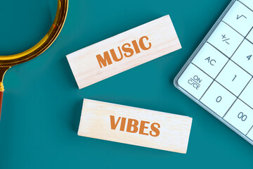 Wooden blocks spelling Music Vibes sit near a magnifying glass and calculator on a Teal surface,...