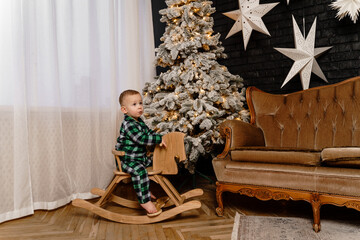 Little boy in green plaid pajamas rides a rocking horse near a Christmas tree