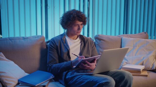 Young Man Studies at Night on Sofa in Neon Light, Holding Notebook and Laptop. Gen Z Study Routine with Calm Productivity and Mood Lighting. Remote Learning, Digital Journaling, Aesthetic Focus.