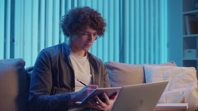 Young Man Studies at Night on Sofa in Neon Light, Holding Notebook and Laptop. Gen Z Study Routine with Calm Productivity and Mood Lighting. Remote Learning, Digital Journaling, Aesthetic Focus.