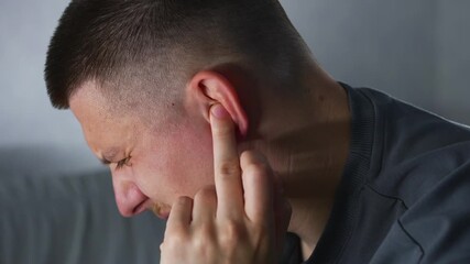 A man sitting on a sofa with an ear infection.
The man touches his sore ear with his hand and suffers from severe pain and aching sensations in his ears.