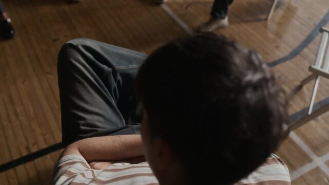 High angle of teenage guy wearing black jeans and striped pullover sitting in front of school psychologist and his schoolmates and listening to advice of counselor at session