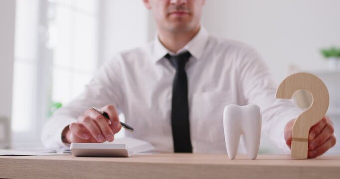 Slow motion video of young business man sitting in office with white tooth and question mark on table calculating costs of expensive dental treatment. Dentistry and stomatology concept. 4k video