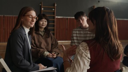Young elegant female counselor putting hand on shoulder of teenage girl explaining her psychological problem while looking for support during session with schoolmates