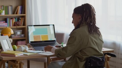 Rear view shot of focused young Caucasian woman with dreadlocks in wheelchair sitting at home office desk in cozy living room, analyzing utility bill and taking notebook to plan monthly budget - Powered by Adobe