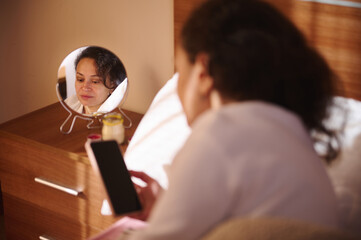 Woman Looks At Her Reflection In A Small Mirror On A Bedroom Dresser While Holding A Phone