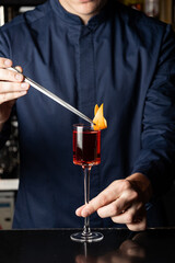 A bartender in a navy uniform carefully garnishes a vibrant red cocktail with an orange peel using tongs. The elegant glass sits on a sleek black bar surface — a stylish snapshot of professional cockt