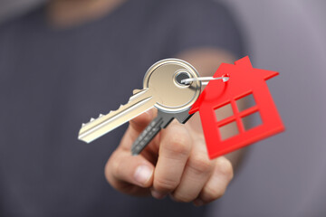 A close-up shot shows a hand presenting house keys with a red house-shaped key chain, conveying real estate ownership and home buying. Focus on