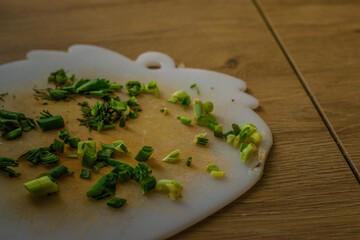 Freshly chopped green onions and herbs scattered on a white plastic cutting board. The board rests on a wooden kitchen surface, hinting at ongoing meal preparation.