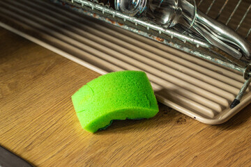 A used green sponge rests on a wooden kitchen counter beside a drying rack. The sponge shows signs of wear, indicating frequent use for dishwashing or cleaning in a domestic setting.