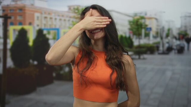 Smiling woman covers eyes with hand on busy urban street with blurred buildings in daylight; shyness happiness.