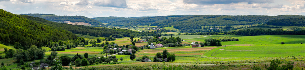 Obraz premium A high-angle panoramic landscape of Pavliv and Kuriany villages near Berezhany, Ternopil Oblast, Ukraine. The houses are nestled in a valley with green fields and surrounded by dense forests.