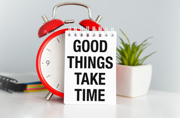 A red alarm clock sits on a table with a white piece of paper that says Good Things Take Time.