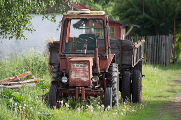 dilapidated red agricultural vehicle positioned near aged farmhouse with overgrown surroundings