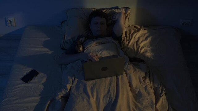 Man lying awake at night using laptop in bed, blue screen light causing insomnia