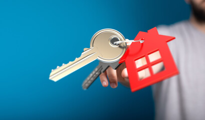 A hand holding house keys, a red house decoration against a blue background. Concept of home ownership, purchase, or real estate.