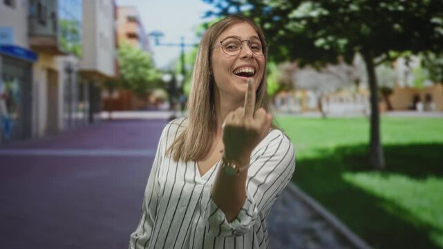 Woman showing middle finger gesture toward camera on a sunlit urban street lined with shops and trees, with blurred buildings and pavement around her; defiance.